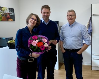 2 Männer und eine Frau bei einer Verabschiedung. Die Frau hat einen Blumenstraß bekommen.